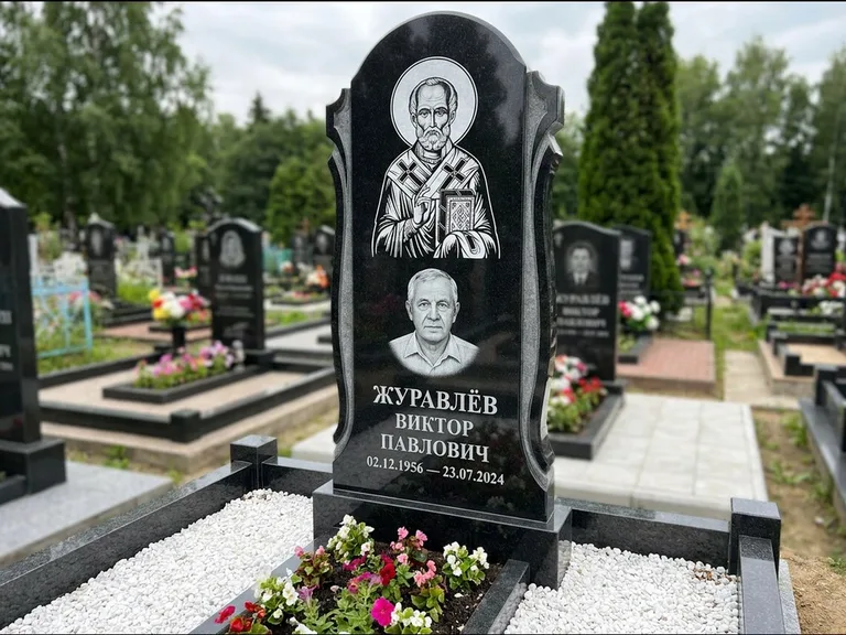 Памятник с гравировкой иконы: стела с образом в полный рост на обороте — гравировка образа Матроны Московской, низкие кусты спиреи в цвету, работа «Лебедева» в Сургуте